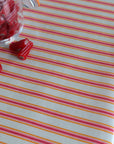 A close-up of a striped oilcloth with alternating pink, orange, and yellow lines on a white base, with a small clear jar of red candy pieces and a few loose red pieces in the upper left.