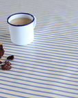A white enamel mug with a blue rim sits on a blue-and-white striped tablecloth, with a half-eaten chocolate cake and crumbs nearby.