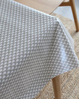A close-up of a light gray tablecloth with a repeating teardrop pattern, draped over a wooden table on a woven jute rug.