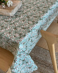 A light green tablecloth with a delicate cherry blossom branch pattern, draped over a wooden dining table with light wood chairs in a cozy, natural setting.