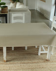 Beige tablecloth on a dining table with white chairs in a room with a rug and plants.