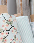 Rolls of fabric with floral and striped patterns in light colors, arranged vertically against a rustic wooden backdrop.
