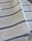 A close-up of a striped tablecloth in cream with thin black and gray horizontal stripes, draped over a table with a small decorative cup visible in the upper left corner.