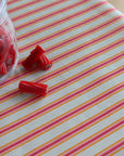 Red licorice candy pieces, some inside a clear glass jar and a few loose, arranged on a white tablecloth with diagonal pink and orange stripes.