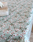 A tablecloth with a delicate floral print of white blossoms on dark branches over a muted sage green background, styled on a table with a rustic wooden block and white flowers.