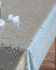 A light blue/green tablecloth with a delicate beige and brown cherry blossom branch pattern, draped over a table with a glass and ice cubes