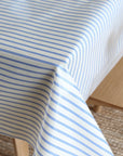 A close-up of a white tablecloth with thin blue horizontal stripes draped over a wooden table in a bright indoor setting.