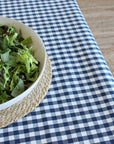 A navy and white gingham tablecloth spread across a wooden table, with a white bowl of mixed green salad resting on a woven beige placemat in the lower-left corner.
