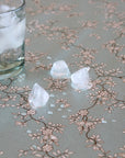 A close-up of a clear glass with ice on a light gray table covered in a delicate beige floral vine pattern, with three ice cubes scattered on the surface.