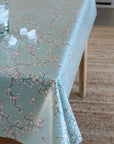 A light blue tablecloth with a delicate white and gray cherry blossom branch pattern, draped over a table in a home setting with a woven rug and a glass of water on top.