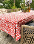 A red tablecloth with a white floral pattern draped over an outdoor wicker dining table on a wooden deck, surrounded by wicker chairs.