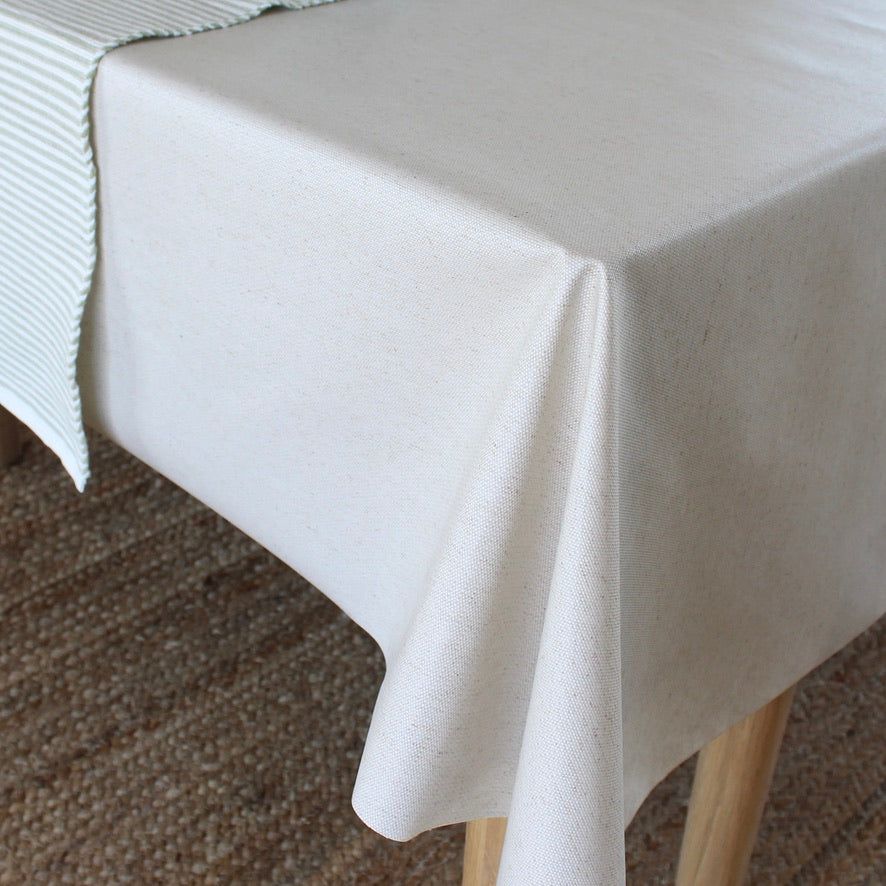 A light beige tablecloth draped over a table with wooden legs, with a striped runner partially visible on the left side over a neutral woven rug.