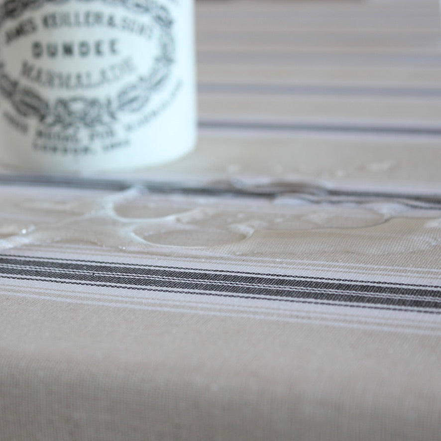 A close-up of a beige tablecloth with thin dark gray stripes and a subtle floral pattern, with a white bottle blurred in the background.