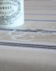 A close-up of a beige tablecloth with thin dark gray stripes and a subtle floral pattern, with a white bottle blurred in the background.