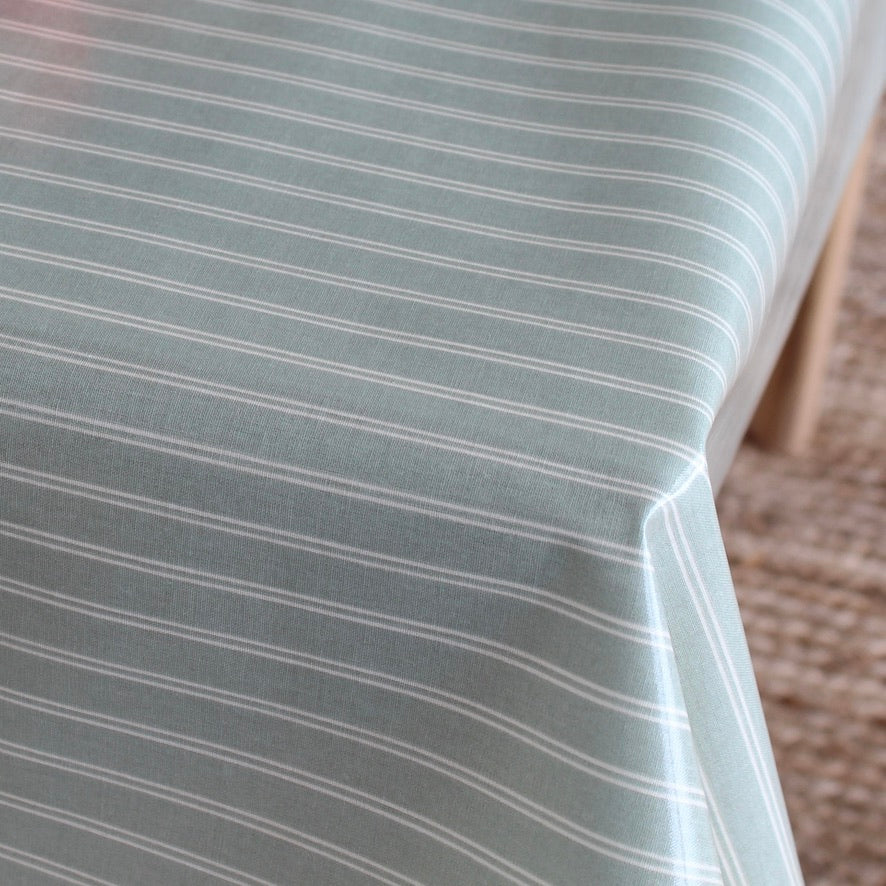 A close-up of a tablecloth with thin white horizontal stripes on a muted sage green background, draped over a table with a woven rug visible in the background.