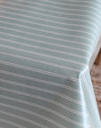 A close-up of a tablecloth with thin white horizontal stripes on a muted sage green background, draped over a table with a woven rug visible in the background.