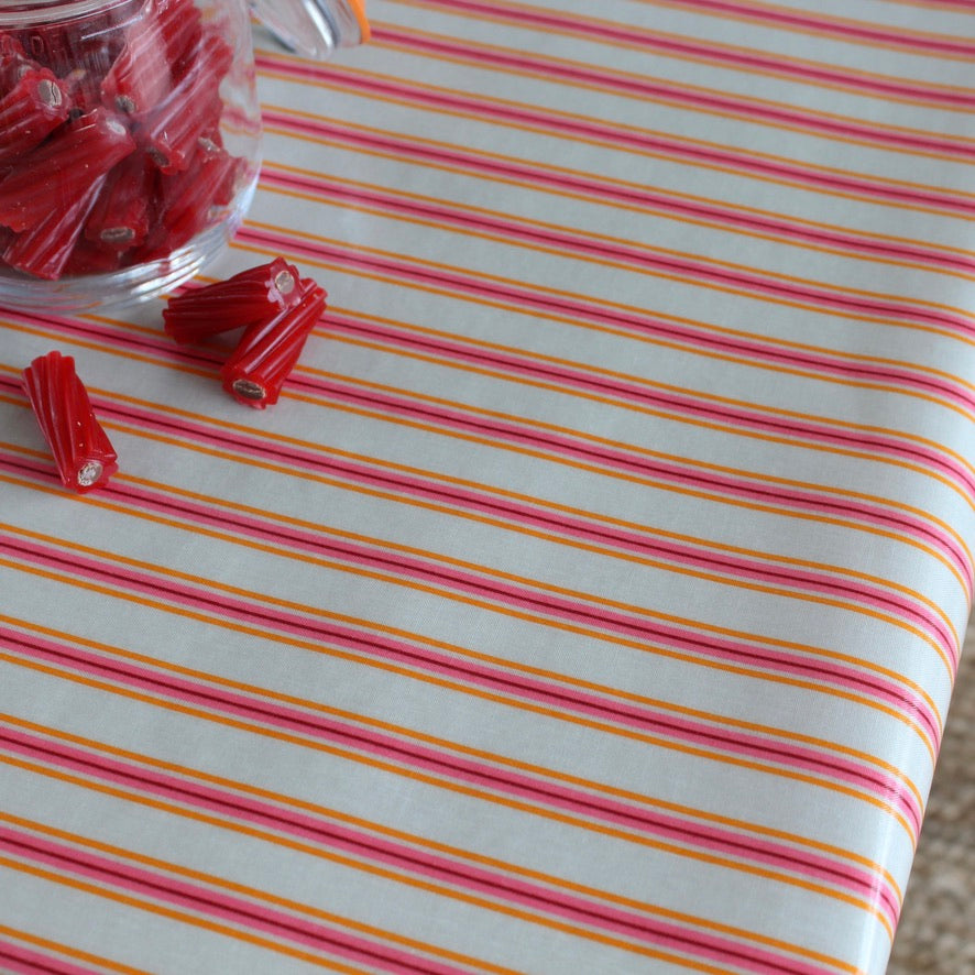 A close-up of a striped oilcloth with alternating pink, orange, and yellow lines on a white base, with a small clear jar of red candy pieces and a few loose red pieces in the upper left.