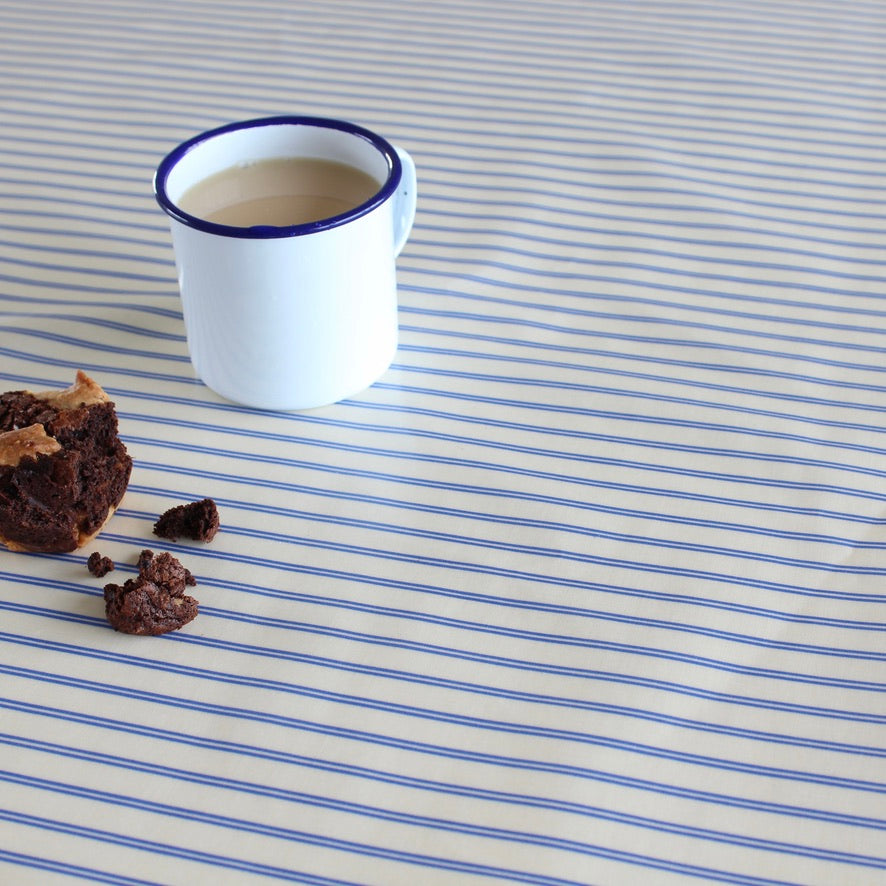 A white enamel mug with a blue rim sits on a blue-and-white striped tablecloth, with a half-eaten chocolate cake and crumbs nearby.