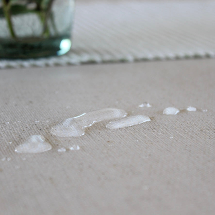 A close-up of a light beige textured oilcloth with a few clear water droplets beaded on top, with a blurred glass vase in the background.