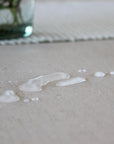 A close-up of a light beige textured oilcloth with a few clear water droplets beaded on top, with a blurred glass vase in the background.