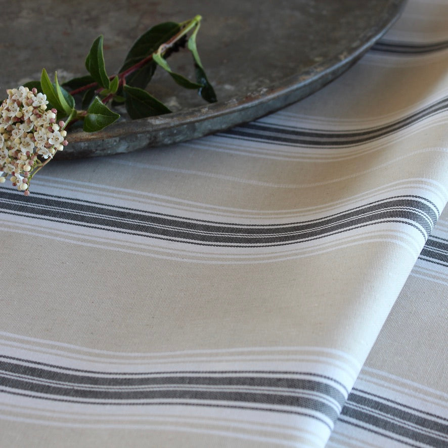 A close-up of a striped tablecloth in warm off-white with dark gray and thin black horizontal stripes. A rustic metal tray and a small green stem with white flowers rest on top as styling props.