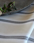 A close-up of a striped tablecloth in warm off-white with dark gray and thin black horizontal stripes. A rustic metal tray and a small green stem with white flowers rest on top as styling props.