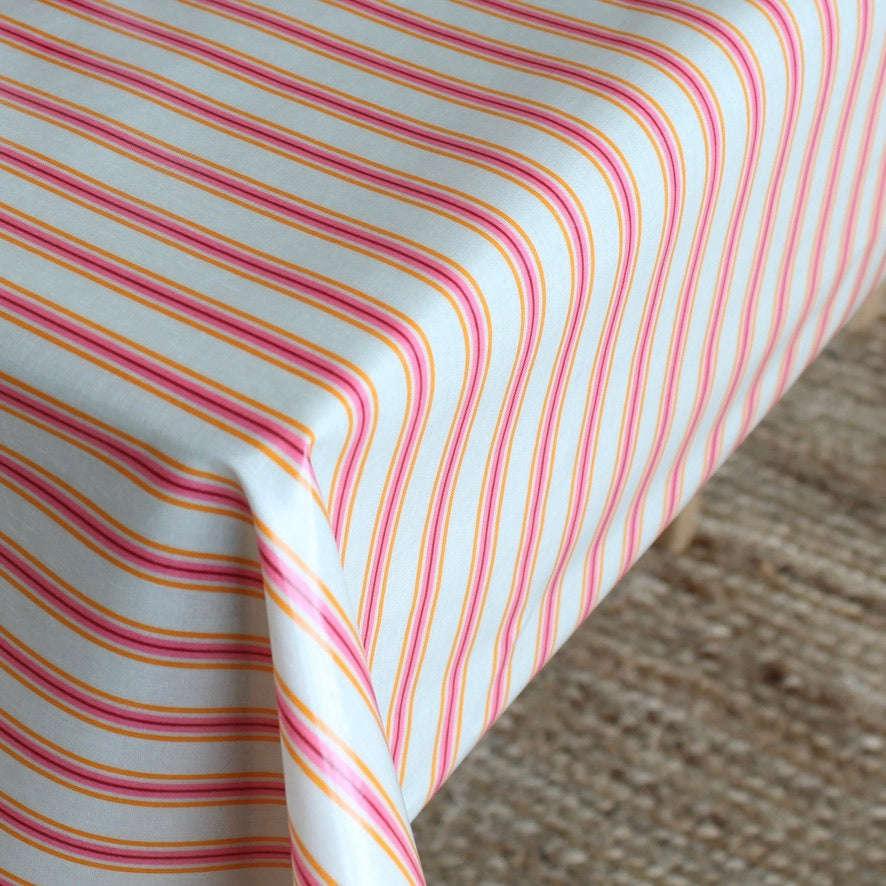 A close-up of a tablecloth with thin pink, orange, and yellow diagonal stripes, draped over the corner of a table with a textured beige rug visible underneath.
