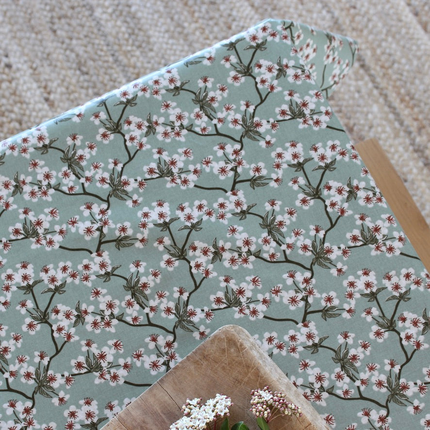An overhead of a tablecloth with a repeating cherry blossom print on a muted sage green background, styled on a wooden surface with small white flowers in the foreground.