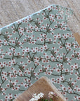 An overhead of a tablecloth with a repeating cherry blossom print on a muted sage green background, styled on a wooden surface with small white flowers in the foreground.
