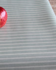 A close-up of a tablecloth with narrow white pinstripes on a muted sage green background, with a red apple placed in the upper left corner.