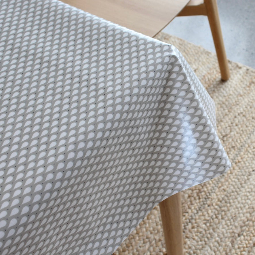 A close-up of a light gray tablecloth with a repeating teardrop pattern, draped over a wooden table on a woven jute rug.