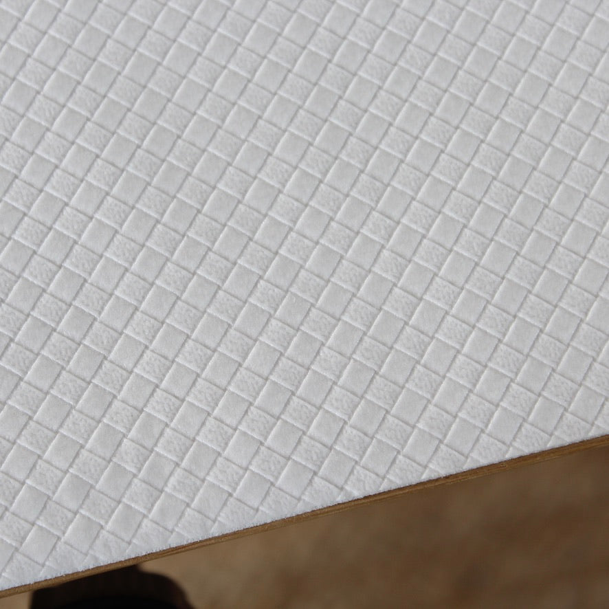 A close-up of a white textured table protector with a repeating small square grid pattern, shown at an angle with a brown edge visible at the bottom.