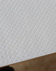 A close-up of a white textured table protector with a repeating small square grid pattern, shown at an angle with a brown edge visible at the bottom.