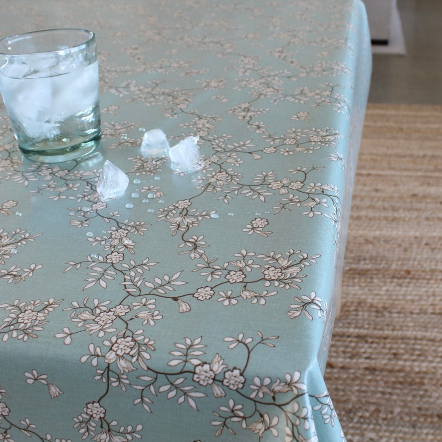A sage green tablecloth with a delicate beige and brown cherry blossom pattern, shown on a table with a glass of water and ice and scattered ice cubes. A woven rug is visible on the right side.