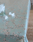 A sage green tablecloth with a delicate beige and brown cherry blossom pattern, shown on a table with a glass of water and ice and scattered ice cubes. A woven rug is visible on the right side.