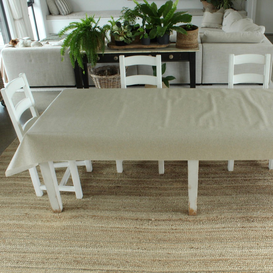Dining room with a long table covered by a beige tablecloth, white chairs, and plants on a wooden table.