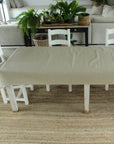 Dining room with a long table covered by a beige tablecloth, white chairs, and plants on a wooden table.