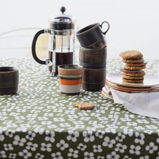 A styled tabletop scene with a green tablecloth covered in a white floral pattern, holding a glass French press, stacked ceramic mugs, a small striped cup, plates with cookies, and a white napkin.