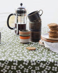A styled tabletop scene with a green tablecloth covered in a white floral pattern, holding a glass French press, stacked ceramic mugs, a small striped cup, plates with cookies, and a white napkin.