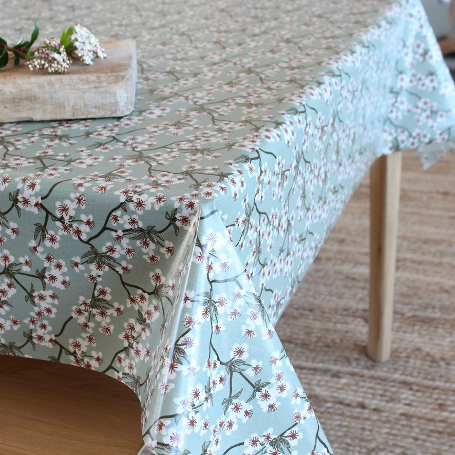 A light sage green tablecloth with a white cherry blossom branch pattern, draped over a wooden table in a softly lit room.