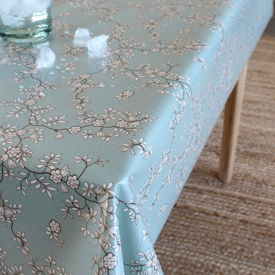 A light blue tablecloth with a delicate white and gray floral branch pattern, draped over a table in a home setting with a woven rug and a glass jar visible.