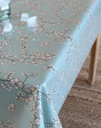 A light blue tablecloth with a delicate white and gray floral branch pattern, draped over a table in a home setting with a woven rug and a glass jar visible.