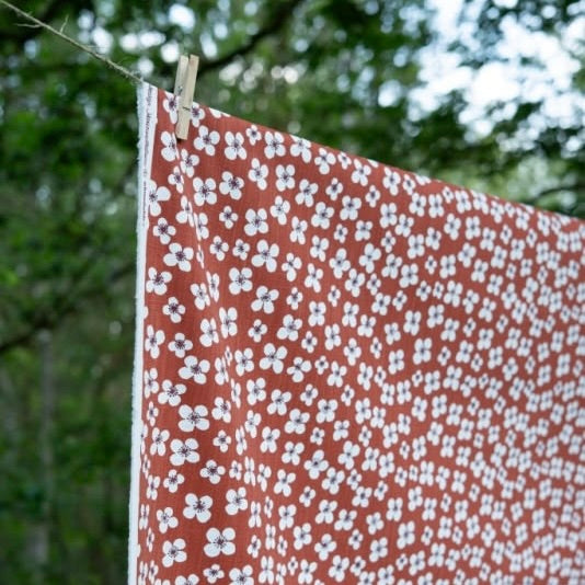 A terracotta orange tablecloth, printed with a white floral block pattern, hanging from a clothesline outdoors with a wooden clothespin.
