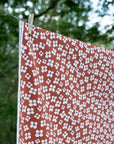 A terracotta orange tablecloth, printed with a white floral block pattern, hanging from a clothesline outdoors with a wooden clothespin.