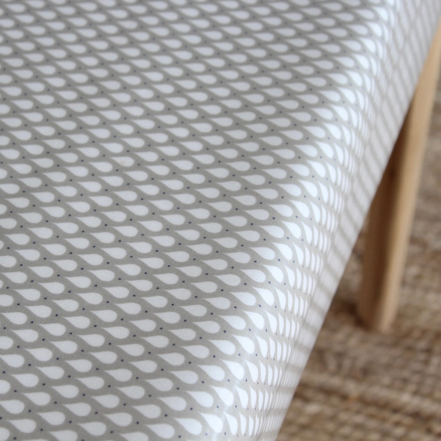 A close-up of a white tablecloth with a repeating gray teardrop pattern, draped over a table with a light wood leg visible on the right side.
