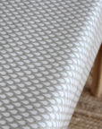 A close-up of a white tablecloth with a repeating gray teardrop pattern, draped over a table with a light wood leg visible on the right side.