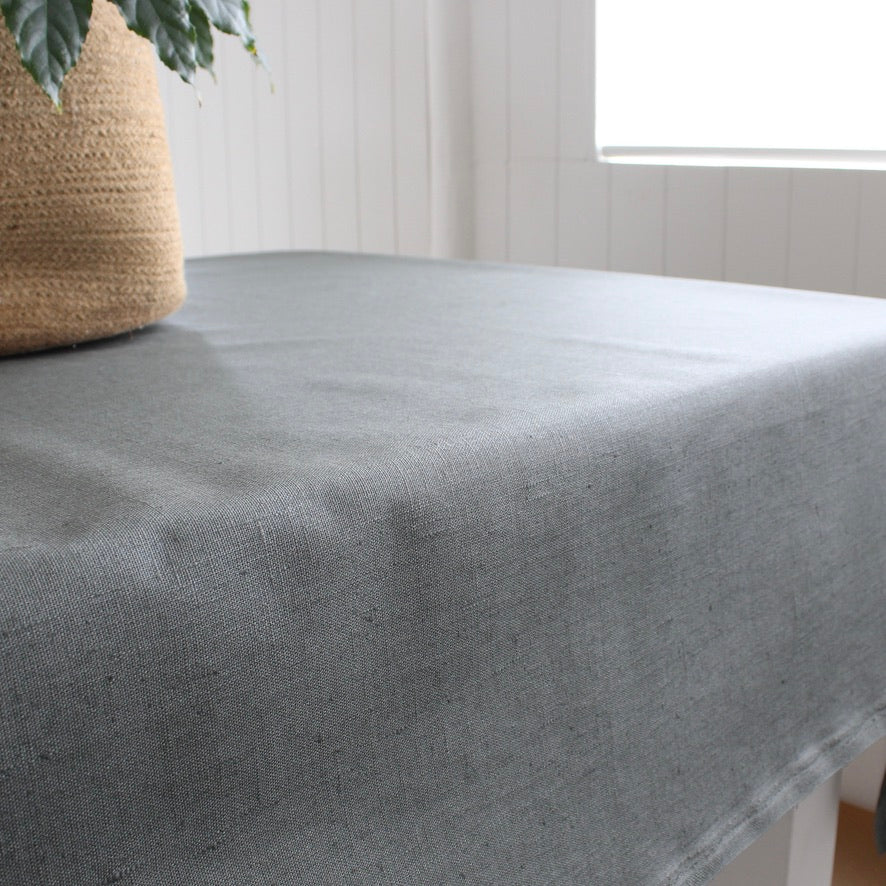Grey green tablecloth on a table with a woven basket and plant in the background