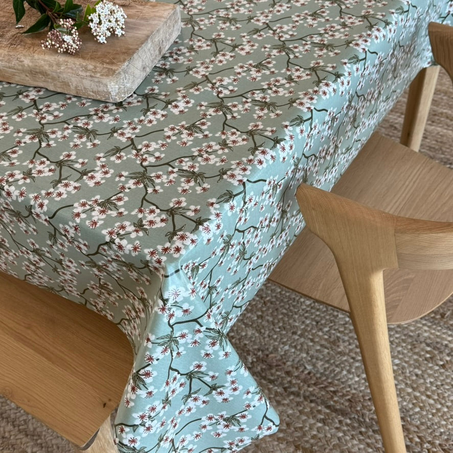 A light green tablecloth with a delicate cherry blossom branch pattern, draped over a wooden dining table with light wood chairs in a cozy, natural setting.