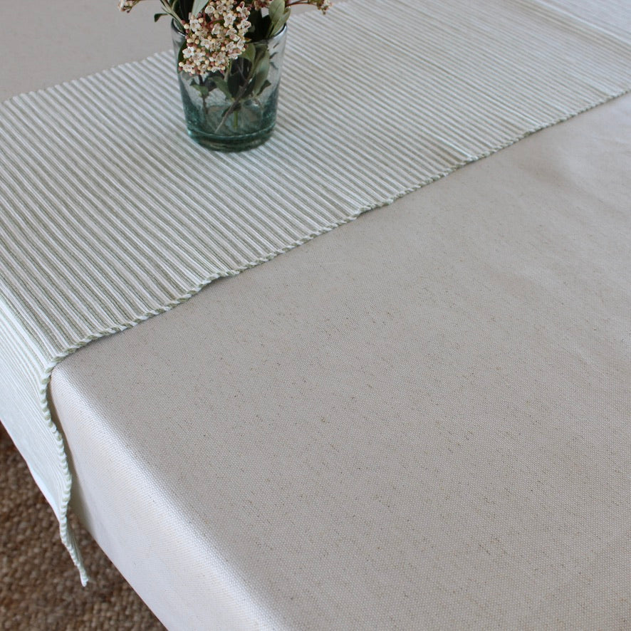 A light beige tablecloth with a matching striped runner laid across a rectangular table, styled with a small glass vase of flowers.