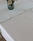 A light beige tablecloth with a matching striped runner laid across a rectangular table, styled with a small glass vase of flowers.